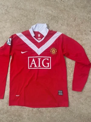 Manchester United långärmad matchtröja - Röd långärmad Manchester United matchtröja från Nike med vitt V-mönster på bröstet, vit krage och AIG-logga på magen. Broderat klubbmärke på vänster bröst och Premier League-märke på ärmen. Perfekt för fotbollsfans som vill sticka ut på planen.