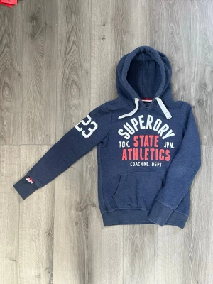 Blå hoodie från Superdry Athletics - Mörkblå hoodie från Superdry med vit och röd text på bröstet och siffran 23 på ärmen🤗 Passar nån med storlek S-Xs🤩 Priset ej hugget i sten därför kan det diskuteras😇