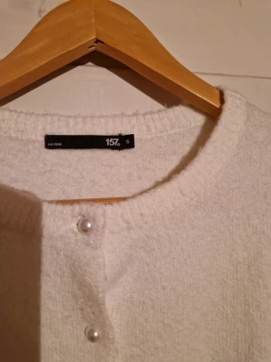 Vit kofta med pärlknappar  - Kortärmad vit kofta från Lager med rund halsringning och pärlknappar framtill. Mjuk stickad känsla och ribbade kanter. Perfekt för en stilren och söt look. Använd några få gånger