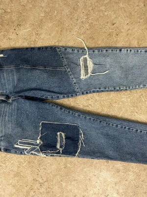 Blå slitna jeans med stora hål - Säljer ett par blå jeans med rejält slitna detaljer och stora hål på benen. Jeansen har en rak passform och är tillverkade i klassisk denim. Perfekta för dig som gillar en edgy och trendig streetstyle-look.