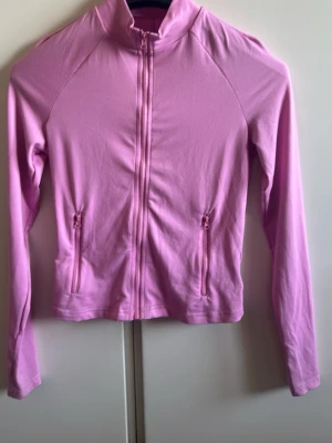Rosa sportjacka med dragkedja - Säljer en rosa sportjacka med hel dragkedja framtill och två fickor med dragkedja. Jackan har lång ärm, hög krage och är tillverkad i ett stretchigt material som passar perfekt för träning eller löpning.