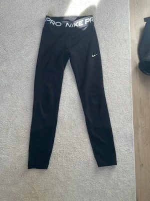 Svarta Nike Pro leggings - Snygga svarta Nike Pro leggings med hög midja och bred resår med Nike Pro-logga. Tight passform och stretchigt material som passar perfekt till träning eller chill. Klassisk vit Nike Swoosh på benet. Materialet känns mjukt och följsamt.