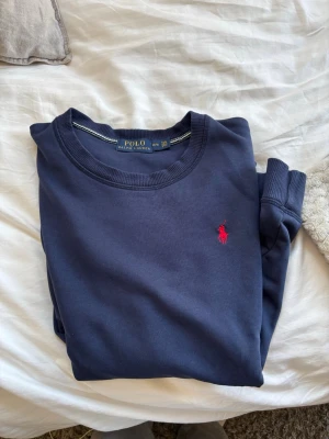 Marinblå sweatshirt från Polo Ralph Lauren - Klassisk marinblå sweatshirt från Polo Ralph Lauren med rund halsringning och den ikoniska röda logotypen broderad på bröstet. Tröjan har långa ärmar och ribbade muddar vid ärmslut och nederkant. Perfekt för en avslappnad och stilren look.