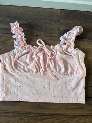 Ljusrosa broderad crop top med volang - Söt ljusrosa crop top med broderade detaljer och volangprydda axelband. Toppen har en liten rosett framtill och är gjord i ett tunt, luftigt tyg med hålmönster. Perfekt för sommaren och ger en romantisk vibe. Xs storlek. 