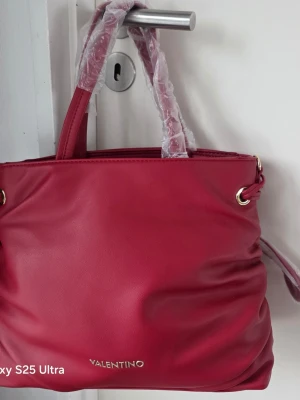 Röd handväska från Valentino - Snygg röd handväska från Valentino med guldfärgade detaljer och dubbla handtag. Väskan är tillverkad i slätt konstläder och har en stilren, mjuk form. Perfekt accessoar för att lyfta din outfit.
