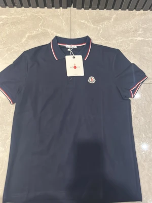 Marinblå polotopp från Moncler - Snygg marinblå polotopp från Moncler med klassisk krage och diskret logotyp på bröstet. Toppens krage och ärmslut har röda och vita detaljer. Materialet är bomull och modellen har korta ärmar och två knappar vid halsen. Logga lite dålig gjort då två stygn missades men loggan sitter fortfarande stabilt därav låga priset. Passar även bra till M 