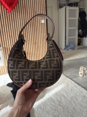 Brun Fendi baguette handväska - Säljer en ikonisk brun baguette handväska från Fendi med det klassiska FF-monogrammet i mörkbrunt och beige. Väskan har en bågformad siluett, kort axelrem i läder och dragkedja upptill. Perfekt accessoar för dig som gillar lyxig och trendig design.