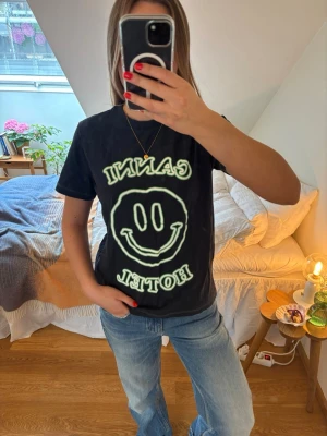 Ganni t-shirt - Cool mörkblå t-shirt från Ganni med stort smiley-tryck och texten 'Ganni Hotel' i vitt/grönt på framsidan. Klassisk passform med korta ärmar och rund halsringning. Perfekt till jeans för en avslappnad look.