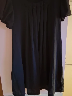 Svart kort klänning med veck - En svart kort klänning med rund halsringning och korta puffärmar. Framtill har klänningen dekorativa veck och små knappar upptill. Enkel och stilren design, perfekt för dig som gillar klassiska plagg.