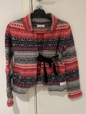 Mönstrad kofta med knytband - Färgglad kofta med mönster i rött, rosa, blått och vitt. Koftan har långa ärmar, bred krage och ett svart knytband i midjan. Materialet ser ut att vara mjukt och stickat, perfekt för kalla dagar. Snygg detalj med fickor på sidan. Den är i odd Molly storlek 1 vilket motsvarar S. Men den är rätt så tajt och sitter bättre som en xxs-xs.