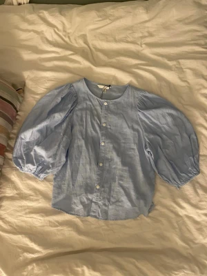 Ljusblå linnemix blus från H&M - Supersöt ljusblå blus från H&M i linnemix med puffiga trekvartsärmar och knappar framtill. Blusen har rund halsringning och en luftig, avslappnad passform som ger en trendig och fräsch look.