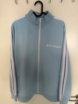 Ljusblå Palm Angels hoodie med dragkedja - Snygg ljusblå hoodie från Palm Angels med vit dragkedja och vita ränder längs ärmarna. 