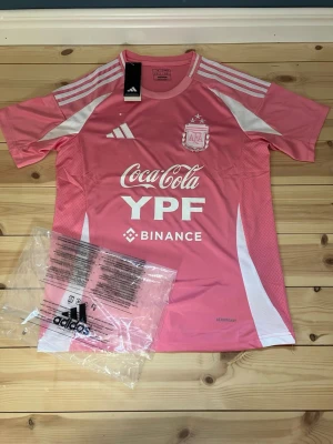 Rosa Argentina tränings t-shirt Adidas - Rosa tränings t-shirt från Adidas med Argentina AFA-logga och sponsorer som Coca-Cola, YPF och Binance. T-shirten har vita detaljer, korta ärmar och klassiska Adidas-ränder på axlarna. Tillverkad i lätt och ventilerande material med Aeroready-teknologi. Väldigt bra kvalitet