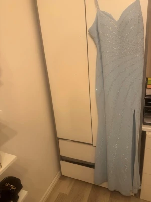 Ljusblå glittrig långklänning - Säljer en elegant ljusblå långklänning med tunna axelband och v-ringning. Klänningen har glittriga detaljer i ett strålande mönster och en hög slits framtill. Perfekt för bal eller festkvällar där du vill sticka ut.