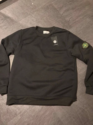 Svart sweatshirt från Stone Island - Svart sweatshirt från Stone Island med klassisk rund hals och ribbade muddar. Tröjan har den ikoniska Stone Island-loggan på vänster ärm och är tillverkad i mjukt bomullsmaterial. Perfekt för en stilren och avslappnad look.
