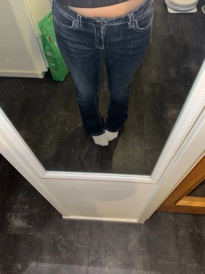 Mörkblå bootcut jeans med brodyr - Säljer ett par mörkblå bootcut jeans med vita kontrastsömmar och snygg broderad ficka bak. Jeansen har mid låg midja och klassisk femficksdesign. Perfekta för dig som gillar detaljer och en avslappnad men trendig look.