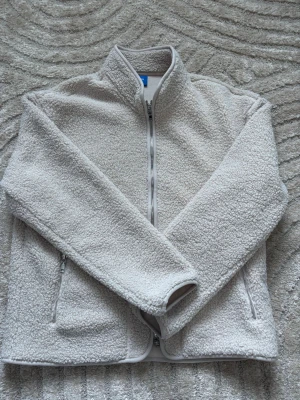 Beige teddyjacka med dragkedja - Mysig beige teddyjacka med hel dragkedja och ståkrage. Jackan har två sidofickor med dragkedja och är tillverkad i fluffigt teddy-material som ger en avslappnad och trendig look. Perfekt för lager-på-lager-stil under kyliga dagar. (HELT OANVÄND KÖPTES FRÅN NLYMAN)