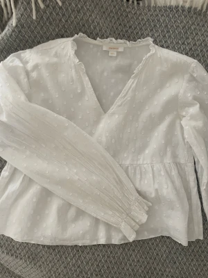 Vit blus med struktur från Gina Tricot - Söt vit blus från Gina Tricot med diskret prickig struktur, v-ringning och volangkant vid halsen. 