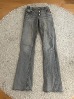 Grå bootcut jeans med knappar - Säljer ett par gråa bootcut jeans med hög midja och tre synliga knappar framtill. Jeansen har klassiska fem fickor och raka ben med lätt utsvängd passform. Perfekta för en avslappnad och trendig look.