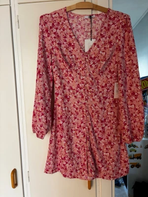 Blommig kortklänning från Nelly - Söt kortklänning från Nelly x NA-KD med rosa och röda blommor på ljus botten. Klänningen har v-ringning, långa ärmar och lätt markerad midja. Perfekt för sommarens alla tillfällen och ger en fräsch och ungdomlig look.