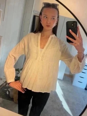 Ljusbeige bohemisk blus med v-ringning - Luftig ljusbeige blus med v-ringning och dekorativa broderade detaljer längs halsen. Blusen har lång ärm med lätt puff och är tillverkad i ett tunt, strukturerat tyg som ger en avslappnad boho-känsla. Perfekt att matcha med jeans eller kjol.