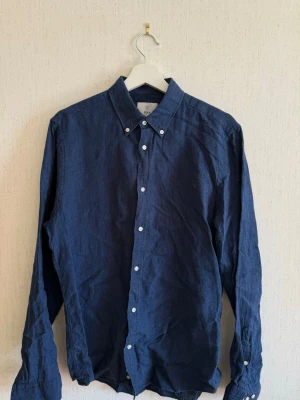 Mörkblå linneskjorta från Bläck - Stilren mörkblå skjorta från Bläck i linnematerial med klassisk krage och vita knappar. Skjortan har lång ärm och en skräddarsydd passform som ger en clean look. Perfekt för dig som gillar enkel men snygg stil.