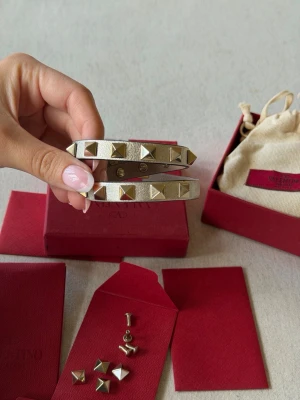 Valentino Rockstud armband i silver/guld - Säljer ett snyggt Valentino Rockstud armband i silverfärgat läder med ikoniska pyramidformade nitar i guld. Armbandet har dubbla remmar och tryckknappsstängning. Perfekt accessoar för att lyfta din outfit med en edgy touch. Kommer med låda och extra nitar🥰