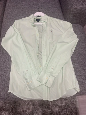 Ljusgrön randig skjorta från GANT - Säljer en ljusgrön randig skjorta från GANT i slim fit. Skjortan har klassisk krage, knappar framtill och långa ärmar. Materialet är bomull och den har en diskret logga broderad på bröstet. Perfekt för dig som gillar stilrena och fräscha plagg.