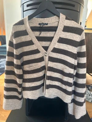 Kofta från Gina Tricot - Mysig kofta från Gina Tricot i beige och bruna breda ränder. Koftan har v-ringning, två framfickor och stängs med en knapp framtill. Ribbstickade muddar och avslut. Perfekt för lager på lager och en chill look.