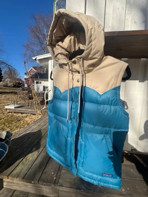 Blå och beige dunväst från Patagonia - Snygg dunväst från Patagonia i blått och beige med stor huva och dragsko. Västen har tryckknappar framtill, två stora fickor och en klassisk outdoor-look. Perfekt för lager-på-lager och riktigt skön att bära under kyliga dagar.
