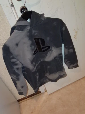 Grå PS5 hoodie med PlayStation-logga (Man) - Säljer en grå hoodie med coolt batikmönster och stor PlayStation-logga på bröstet. PS5-tryck på ryggen. Hoodien har huva och en stor magficka. Perfekt för gamers som vill ha något snyggt och bekvämt. Materialet känns mjukt och skönt.