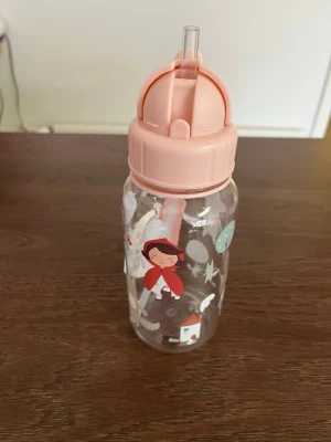 Rosa vattenflaska med sagomotiv - Söt vattenflaska i plast med rosa lock och sugrör. Flaskan är dekorerad med motiv av rödluvan, ballonger och små hus. Perfekt att ha med sig till skolan eller träningen. Rymmer ca 3-5 dl och har praktiskt bärhandtag på locket.