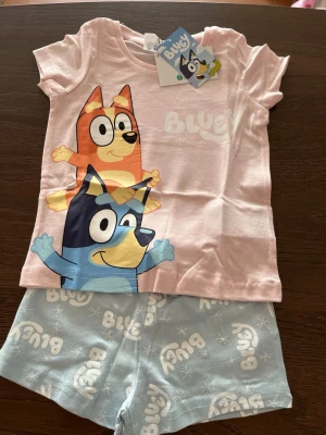 Bluey pyjamas med tryck - Söt pyjamas från Bluey med kortärmad ljusrosa topp och ljusblå shorts. Toppen har ett färgglatt tryck med Bluey och Bingo, medan shortsen har vit Bluey-logga och stjärnmönster. Perfekt för mysiga kvällar och loungewear.