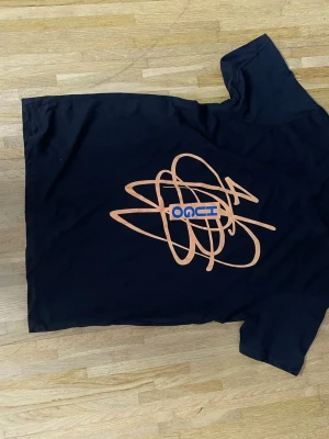 Svart t-shirt med orange grafiskt tryck - Cool svart t-shirt med ett stort orange grafiskt tryck och blå text på framsidan. T-shirten har klassisk passform och korta ärmar, perfekt för en avslappnad stil.