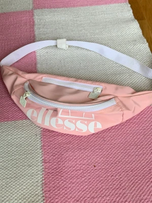 Rosa Ellesse magväska - Snygg rosa magväska från Ellesse med vit dragkedja och justerbart vitt band. Väskan har ett stort Ellesse-tryck framtill och är tillverkad i ett lätt, tåligt material. Perfekt för att bära det viktigaste när du är på språng. Svart/grå fläck på framsidan men annars i fint skick.