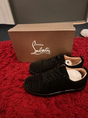 Svarta Christian Louboutin sneakers - Svarta sneakers från Christian Louboutin med klassisk röd sula och svarta nitar på tån. Skorna har lågt skaft, snörning och är tillverkade i mocka och läder. Perfekta för dig som vill sticka ut med en lyxig och edgy stil.