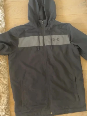 Svart hoodie från Under Armour - Snygg svart hoodie från Under Armour med dragkedja och huva. Har en grå panel över bröstet med logga och två fickor framtill. Tillverkad i ett lätt och ventilerande material med meshfoder. Perfekt för en sportig och avslappnad stil. Storlek S passar M och S