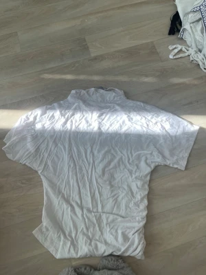 Vit t-shirt med hög krage - Vit t-shirt med hög krage och korta ärmar. Materialet känns mjukt och lätt, perfekt för en chill look. Enkel design utan mönster eller detaljer, passar dig som gillar clean och stilrent.