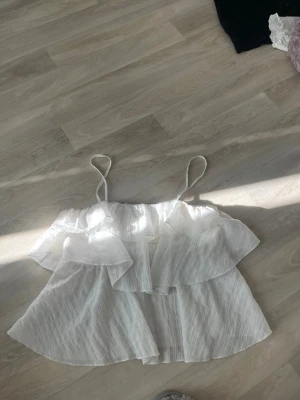 Vit volang linnetopp med tunna band - Säljer en vit linnetopp med tunna axelband och volangdetaljer. Toppen är ärmlös och har en luftig, flowy passform som känns perfekt för varma dagar. Materialet är lätt och har en subtil struktur. Passar dig som gillar en feminin och avslappnad stil.