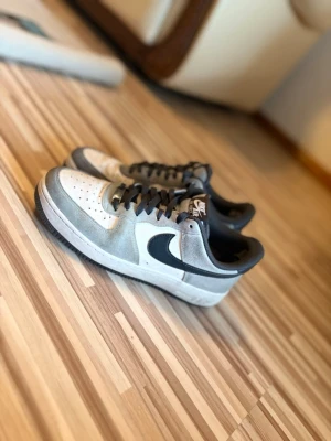 Nike Air Force 1 grå och vita sneakers - Säljer ett par Nike Air Force 1 sneakers i grått och vitt med svart swoosh. Skorna har klassisk låg profil, perforerad tå och svarta skosnören. Materialet är en mix av läder och mocka, perfekt för dig som gillar stilrena och tidlösa sneakers.