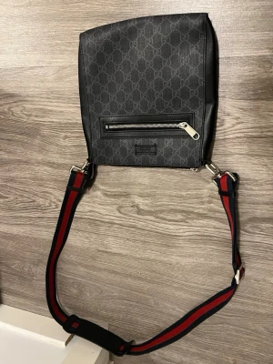 Svart axelväska gg - Cool svart axelväska med klassiskt GG-mönster över hela väskan. Väskan har en silverfärgad dragkedja på framsidan och en justerbar axelrem i svart och rött. Materialet är slitstark canvas med läderdetaljer. Perfekt för dig som vill ha en stilren och trendig väska.