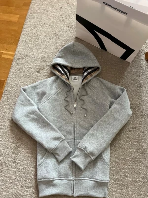 Grå zip-hoodie från Burberry - Snygg grå hoodie från Burberry med dragkedja och klassiskt rutigt foder i huvan. Broderad logga på bröstet och ribbade muddar. Perfekt för en stilren och avslappnad look. Materialet är mjuk bomullsblandning.