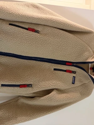 Beige fleecejacka från Patagonia - Säljer en beige fleecejacka från Patagonia med blå dragkedjor och röda detaljer. Jackan har flera fickor med dragkedja och är tillverkad i mjukt, värmande fleecematerial. Perfekt för lager-på-lager och outdoor vibes. Storlek medium, pris kan diskuteras. Hör av dig !