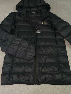 Svart pufferjacka från EA7 Armani - Snygg svart pufferjacka från EA7 Emporio Armani med huva och dragkedja framtill. Jackan har quiltad design, två sidofickor med dragkedja och logga på bröstet. Perfekt för kalla dagar och riktigt trendig stil.