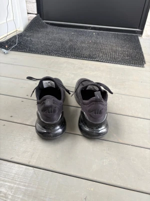Svarta Nike Air Max 270 sneakers - Säljer ett par helsvarta Nike Air Max 270 sneakers med mesh-ovandel och synlig Air-sula. Skorna har klassisk Nike-logga på hälen och snörning framtill. Perfekta för dig som gillar sportig och clean stil.