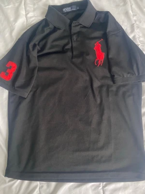Svart Polotröja från Ralph Lauren - Svart pikétröja från Ralph Lauren med röd broderad logga på bröstet och en stor röd siffra 3 på ärmen. Klassisk krage och knäppning framtill. Perfekt för en stilren och sportig look. Storleken är L men passar M
