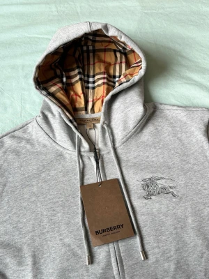 Grå zip hoodie från Burberry - Snygg grå zip hoodie från Burberry med klassiskt rutigt foder i huvan och broderad logga på bröstet. Hoodien har dragsko i huvan, fickor fram och metalländar på snörena. Perfekt för dig som gillar exklusiv streetwear och vill ha något som sticker ut. Passar bättre till storlek s 