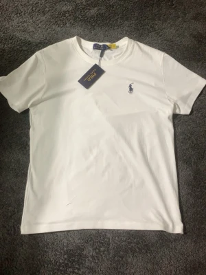 Vit t-shirt från Polo Ralph Lauren - Vit t-shirt från Polo Ralph Lauren med klassisk rund hals och korta ärmar. T-shirten har det ikoniska broderade Polo-loggan i mörkblått på bröstet. Tillverkad i mjuk bomull för en skön känsla och stilren look.