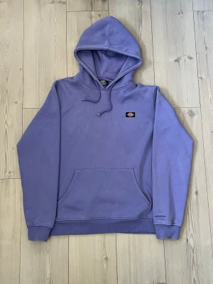 Lila hoodie från Dickies - Snygg lila hoodie från Dickies med klassisk huva och magficka. Diskret Dickies-logga på bröstet och broderad detalj vid ärmslutet. Mjuk bomullsmix som är skön att ha på sig. Perfekt för en avslappnad streetstyle-look.