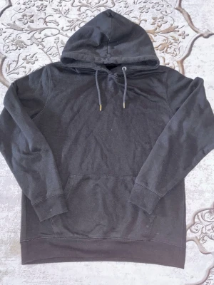 Svart hoodie från 157 med huva - En stilren svart hoodie från 157 med klassisk huva och dragsnören. Hoodien har en stor magficka och ribbade muddar vid ärmslut och nederkant. Perfekt för en avslappnad och cool look. Materialet känns mjukt och bekvämt mot huden.